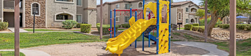 A playground with a yellow slide in the middle of a grassy area.
