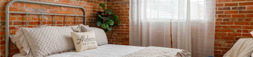 A bed with a white comforter and pillows with the word Alabama on them sits in a room with a brick wall and a window with white curtains.