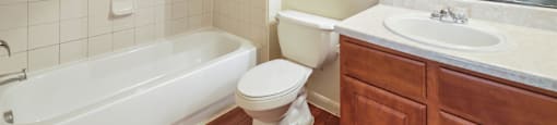 A white toilet sits in a bathroom with a white tub and wooden cabinets.