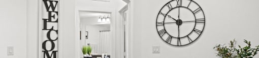 A dining room with a black table and chairs, a clock on the wall, and a "Welcome" sign on the door.