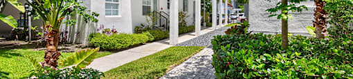 A white building with a black railing is surrounded by green plants.