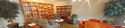 A room with a long bookshelf, a couch, a chair, a table, and a potted plant.