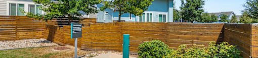 A green post box sits in the middle of a gravel area in front of a wooden fence.