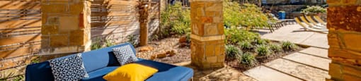 A blue couch with a yellow pillow is under a wooden pergola.