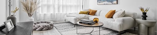 A modern living room with a white sofa, a grey coffee table, and a large window with white curtains.