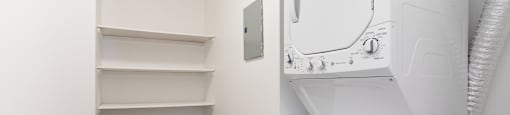 A white dryer is in a small room with a white wall and a white shelving unit.