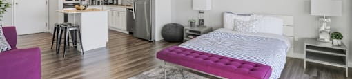 A bedroom with a purple bed and a kitchen in the background.