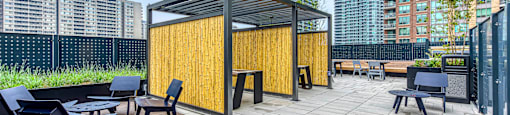 A wooden pavilion with a black roof is surrounded by black chairs and tables on a wooden deck.