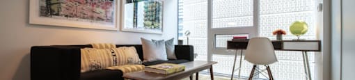 A living room with a black couch, a table with a lamp, a chair, and a window with blinds.
