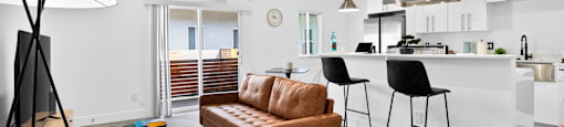 A modern living room with a brown leather sofa and a white kitchen in the background.