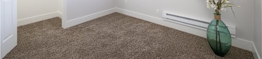 A room with a carpeted floor, a window with blinds, and a green vase with white flowers.