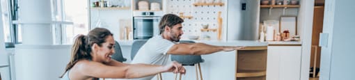 A woman and a man are exercising together with a small dog in a kitchen.
