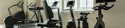 A row of treadmills, stationary bikes, and elliptical machines are lined up in a row in a brightly lit room.