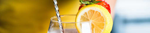 A person in a yellow shirt is stirring a pink drink with a lemon slice and a strawberry on the rim of the glass.