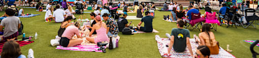 A large group of people are sitting on the grass in front of a building with a sign that reads "Pearl".
