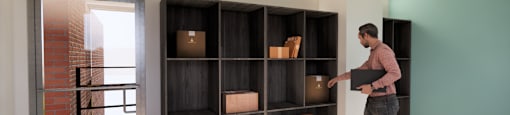 A man is carrying a box into a room with a large shelf.