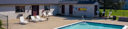 A swimming pool in front of a building with a white fence and lawn chairs.