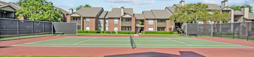 A tennis court is surrounded by a green lawn and apartment buildings.