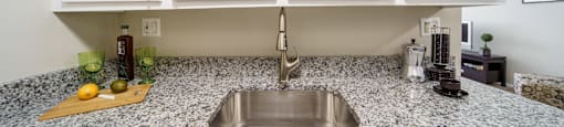 Stainless steel sink with white upper cabinets and  grey and white stone counters.