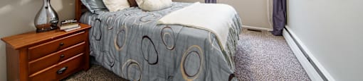 Bedroom with plush carpeting, bed with blue bedding, and wooded nightstand with lamp on top.