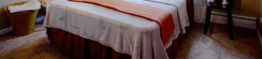 Bed against window with white curtains on each side of window.  Two-tone beige walls with white accent trim.  Striped orange bedding, beige carpet and photo on left wall.  Basket under photo and table to right of bedside.