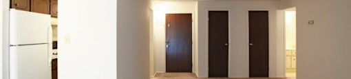 Living area with beige carpeting, dark brown doors to coat closet and exit of apartment, looking onto kitchen