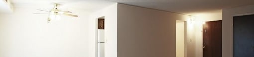 Living area with beige carpeting, ceiling fan, and doorway leading to galley style kitchen, door to right leading outside of apartment.
