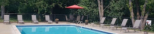 Rectangle pool with brown lawn chaairs, table with 4 chairs and red umbrella in back corner