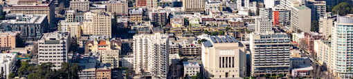 A cityscape with a mix of modern and older buildings, a bridge in the distance, and a mountain range in the far background.