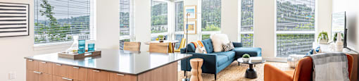 A large living room with floor to ceiling windows, dark wood plank flooring, breakfast bar, and an abundance of natural light.