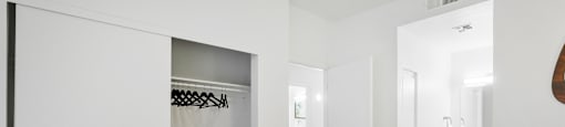 A white bed with a white pillow is in the foreground of a room with a white wardrobe and a white door.