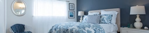 Sienna apts bedroom with a dark teal wall a bed placed int he middle of two white nightstands with blue flower bedspread.