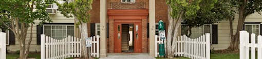 Apartments for Rent in Van Nuys CA - Colonial Manor - Exterior View Of Apartment Complex Entrance Featuring Stone Walkway, Lush Green Grass and Trees, And White Picket Fence