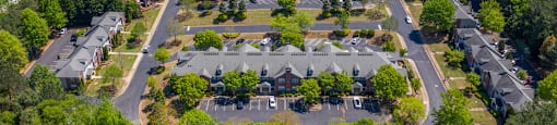Aerial view of a large apartment community surrounded by lush greenery and wooded areas. The complex features multiple rows of townhome-style buildings with brick exteriors, neatly arranged parking lots, and well-maintained landscaping along winding roads