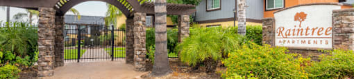 A brick pathway leads to a gate with an archway and a sign that says Raintree Apartments.