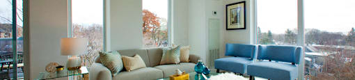 Living room surrounded by large windows overlooking the nearby scenery. There are two fluffy white stools and two blue chairs in the center of the room