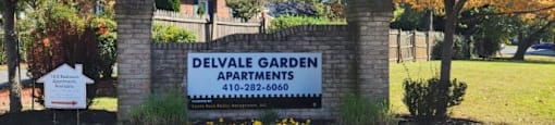 A sign for Delvale Garden Apartments is surrounded by flowers and trees.