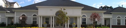A large white house with a grey roof and a circular flower bed in front.