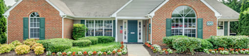 The Leasing Office building is mainly brick with a small amount of gray siding and forest green shutters and front door. There are flower beds on either side of the sidewalk leading to the front door.