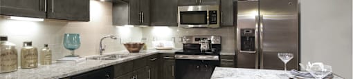 An open plan kitchen with white granite countertops, overhead and under the counter dark wood cabinetry, steel appliances, and built-in wineracks