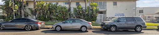 Three cars are parked in a lot with a building and palm trees in the background.