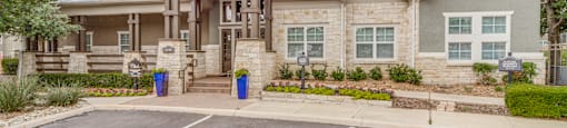 Three Bedroom Apartments in San Antonio TX - Club at Stone Oak - Large Exterior of a Building with Stone Walls, Tan Coloring, Landscaped Plant Beds, and Parking