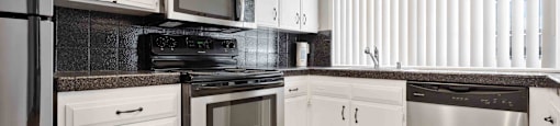 Gilroy, CA, Apartments for Rent - Mission Park - Kitchen with Tan Tile Flooring, Stainless Steel Appliances, Black Granite Countertops, Black Textured Tile Backsplash, White Cabinetry, and Large Window with Vertical Blinds.