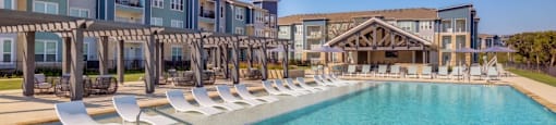 Apartments for Rent in Georgetown Texas - WIlliamson at the Overlook - Sparkling Pool Surrounded by Poolside and Submerged Lounge Seating, with Tables and Chairs Under Pergolas, and View of Apartment Buildings in the Background