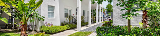 A white building with a black railing is surrounded by green plants.
