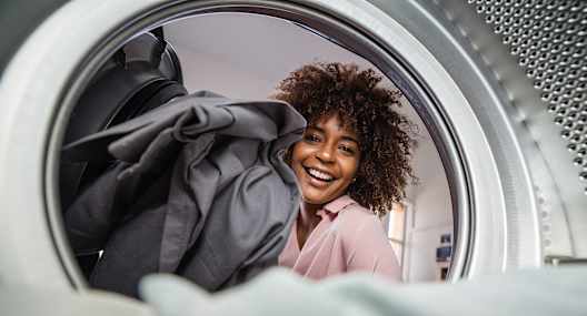 A woman is smiling while looking at clothes inside a washing machine.