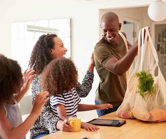 a family of four is gathered around a kitchen table with a grocery bag of vegetables
