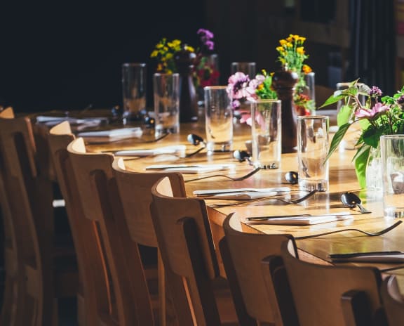Long brown wooded tabled with glass vases filled with flowers and a set table. Wood chairs that match the table are tucked under it. 