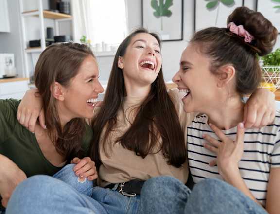 Three women are laughing and hugging each other in a kitchen.