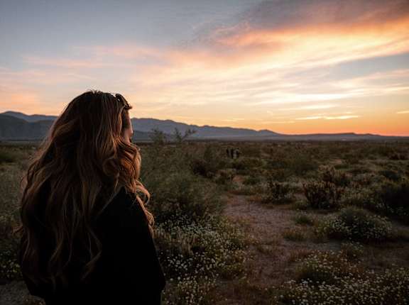 A woman with long hair stands in a field at sunset.
